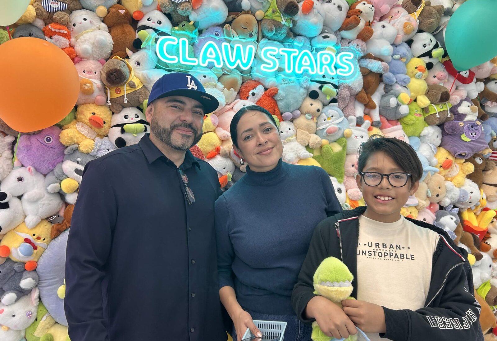 Family at the neon Claw Stars plushie wall, kid clutching a freshly won green plushie